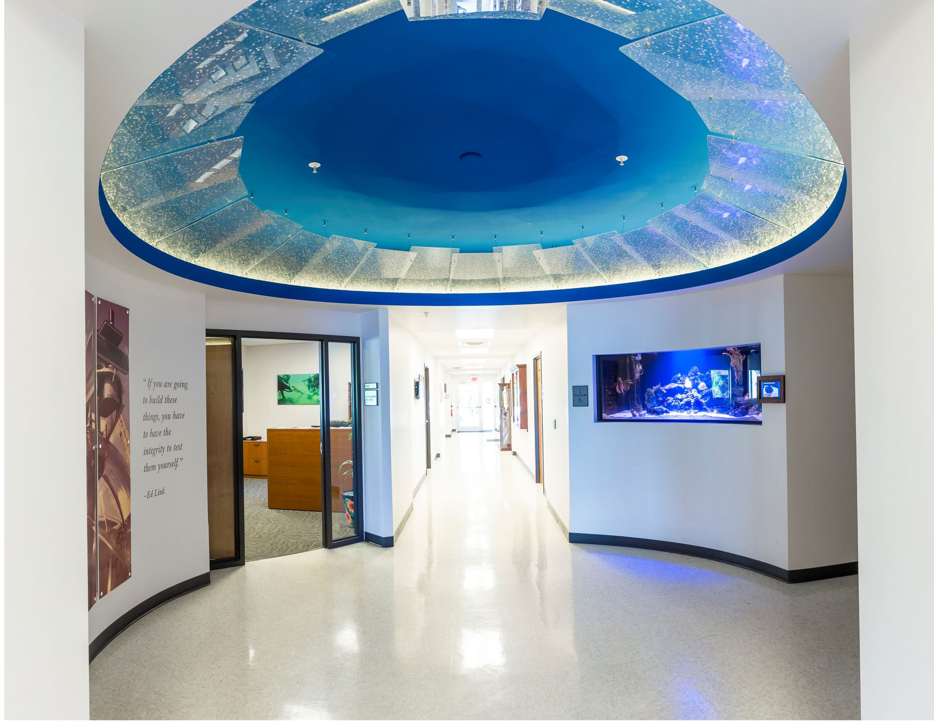 Modern hallway in a university building featuring a large, illuminated blue overhead structure and an embedded aquarium display in the wall, with offices and motivational quotes along the corridor